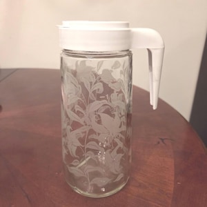 May include: A clear glass pitcher with a white plastic lid and handle. The pitcher features an etched floral design. The handle is attached to the side of the pitcher. The pitcher is sitting on a wooden surface.