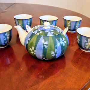 May include: A ceramic tea set with a teapot and five matching cups. The teapot and cups feature a design of vertical stripes in blue and green, with white floral accents. The set is arranged on a wooden table.