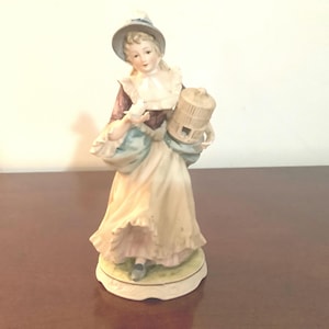 May include: A porcelain figurine of a woman in a long dress and hat, holding a birdcage and a bag with a bird. The figurine has a cream-colored dress, a purple bodice, and a light blue bag. The base is oval-shaped with a green and yellow design.