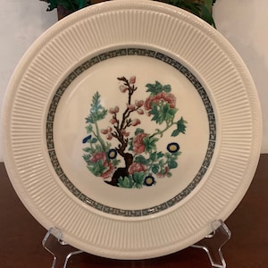 May include: Cream-colored decorative plate with a floral design. The center features a detailed illustration of a flowering tree with pink and green blossoms. The plate has a ribbed border and a dark green and black patterned band.