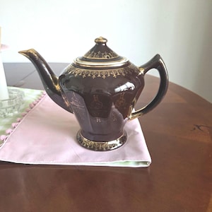 May include: A brown ceramic teapot with gold trim and accents. The teapot has a curved spout, a rounded handle, and a lid with a decorative finial. The teapot is sitting on a pink cloth.