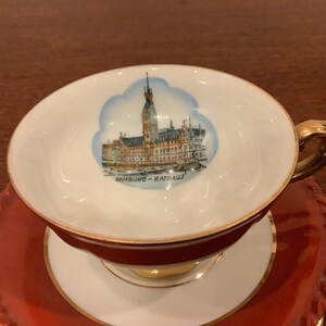 Może przedstawiać: Zestaw filiżanki i spodka do herbaty w stylu vintage. Filiżanka przedstawia szczeg&oacute;łową ilustrację Ratusza w Hamburgu z napisem &bdquo;HAMBURG - RATHAUS&rdquo; poniżej. Filiżanka i spodek są gł&oacute;wnie białe i czerwone ze złotymi akcentami.