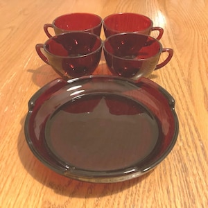 May include: A set of four ruby red glass teacups with handles and a matching round serving dish. The cups and dish are arranged on a light brown wooden surface. The cups are empty and have a simple, classic design.