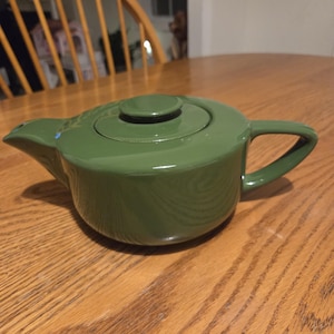 May include: A green ceramic teapot with a lid and handle. The teapot is a solid, glossy green color and sits on a wooden table. The teapot is designed for brewing tea.