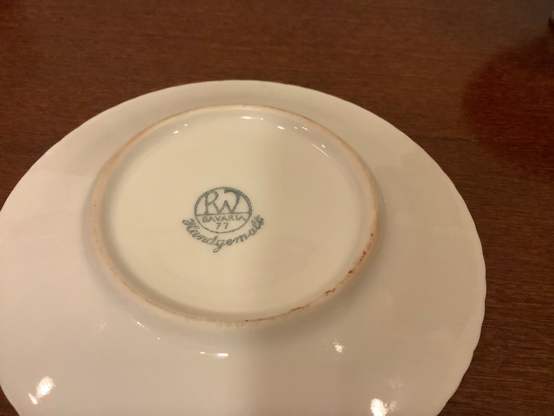 Może przedstawiać: Biały talerz porcelanowy z falistym brzegiem. Sp&oacute;d zdobi niebiesko-białe logo z napisem "RW Bavaria 77 Handgemalt."