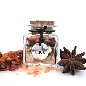 May include: A small glass jar filled with pink Himalayan salt, dried flowers, and star anise. A silver charm with the word "Luck" hangs from the jar.