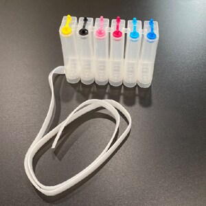 Puede incluir: Un juego de siete cartuchos de tinta de pl&aacute;stico transparente con tapas de colores. Los cartuchos est&aacute;n dispuestos en fila y conectados por una cuerda blanca.
