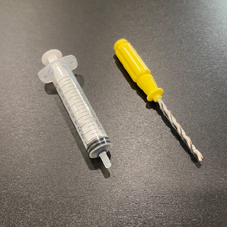 Puede incluir: Una jeringa de pl&aacute;stico transparente con una etiqueta en blanco y negro que dice "DISCARD AFTER USE" y una broca de pl&aacute;stico amarilla con una broca de metal plateado.
