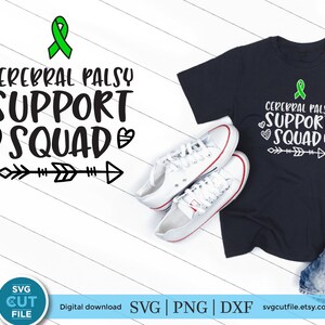 May include: A black t-shirt with a green ribbon and the text "Cerebral Palsy Support Squad" in white. The text is surrounded by small white hearts and arrows. The t-shirt is on a white wooden surface with a pair of white sneakers.