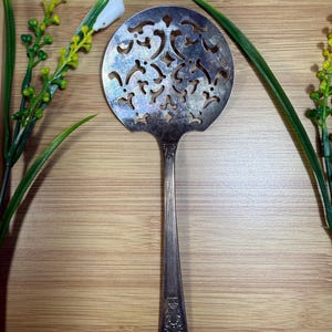 May include: A silver-plated serving spoon with a wide, circular, perforated bowl featuring an intricate cut-out pattern. The handle is slender and straight, decorated with embossed scrollwork at the base, showing signs of natural tarnish.