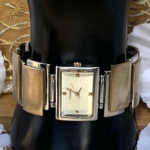 May include: A Fossil wristwatch featuring a rectangular silver-tone case and a cream-colored dial with a sunburst pattern. The watch has a wide, segmented bracelet band that alternates between polished silver-tone and brushed gold-tone metal links.