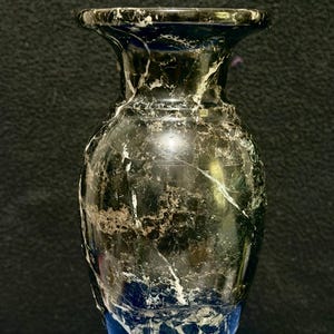 May include: A polished stone vase featuring a dark green and black marbled pattern with prominent white veining. The vase has a flared rim, a rounded body, and a tiered circular base, showcasing a smooth, reflective surface throughout the piece.