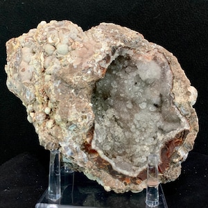 May include: A geode, a hollow rock formation, is displayed on a clear stand. The exterior is a mix of beige, brown, and tan, with a rough, textured surface. The interior reveals a cavity filled with sparkling, translucent crystals.