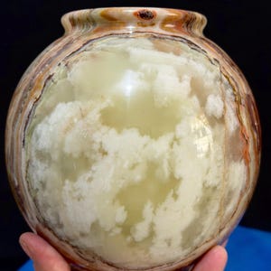 May include: A round, decorative vase with a wide body and a narrow neck. The vase is made of a translucent stone with a cloudy, white and pale green pattern. The rim has brown and orange bands.