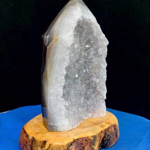 May include: A large, geode crystal with a pointed top, set on a natural wood base. The crystal features a white interior with sparkling formations and a gray and brown outer layer. The wood base is a light brown color with a rough, natural edge.