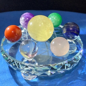 May include: Seven crystal spheres of various colors, including red, orange, yellow, green, blue, purple, and pink, are arranged on a clear crystal base. The spheres are arranged in a circle around the base.