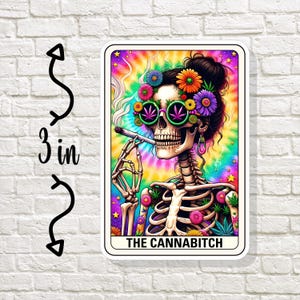 May include: A colorful tarot card with a skeleton wearing sunglasses and a flower crown. The card is titled "The Cannabitch".