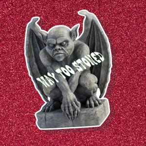 May include: A gray gargoyle statue with large wings, perched on a stone base. The statue has a stern expression and the text "WAY TOO STONED" is printed across it. The background is a sparkly red.