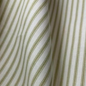 May include: Close-up of a fabric with vertical stripes. The fabric has a white background with evenly spaced, narrow stripes in a muted green color. The fabric is slightly folded, showing the texture and weave of the material.