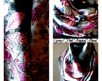 Op de afbeelding: Een grijze en roze tropische bloemen infinity sjaal. De sjaal heeft een herhalend patroon van roze bladeren en zwarte accenten op een grijze achtergrond. De tekst "Grey/Pink Tropical Floral" is zichtbaar.