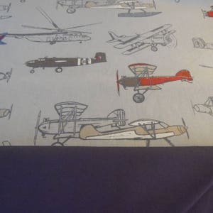 May include: A gray and white fabric with a repeating pattern of various airplanes in different colors and styles. The airplanes are all in flight and some have propellers.