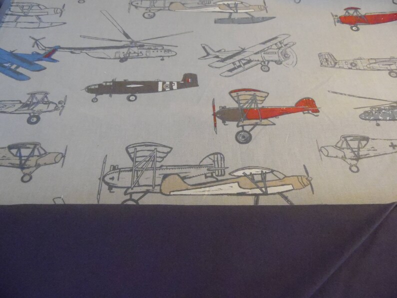 May include: A gray and white fabric with a repeating pattern of various airplanes in different colors and styles. The airplanes are all in flight and some have propellers.