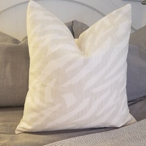 Puede incluir: Una almohada blanca con un sutil estampado de cebra en gris claro. La almohada est&aacute; sobre una colcha gris y blanca.