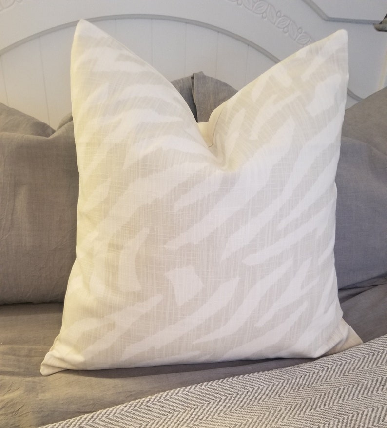 Puede incluir: Una almohada blanca con un sutil estampado de cebra en gris claro. La almohada est&aacute; sobre una colcha gris y blanca.