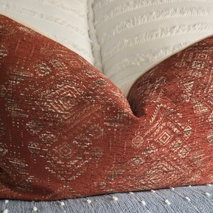 May include: A rust-colored throw pillow with a geometric pattern in a cream color. The pillow is made of a textured fabric.