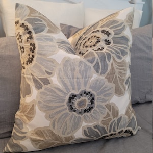 May include: A decorative pillow with a large floral pattern in shades of gray, beige, and black. The pillow is placed on a bed with gray linens, creating a neutral and calming aesthetic. The pillow's design features large, stylized flowers.