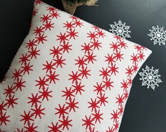 May include: A square, cream-coloured cushion featuring a repeating pattern of red starbursts. The cushion is part of a festive theme, with a small Christmas tree and snowflake decorations in the background.
