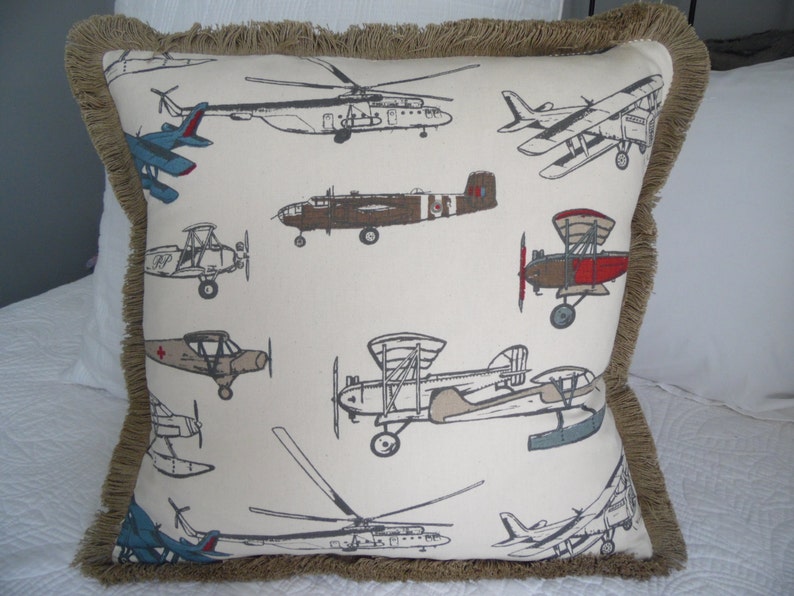 May include: A beige pillow with a brown fringe border features a repeating pattern of vintage airplanes in various colors and styles.