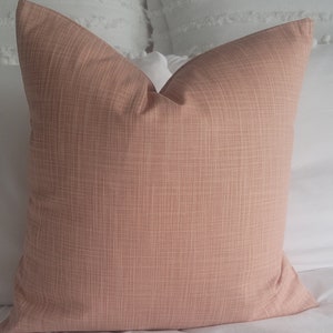 May include: A soft, blush pink throw pillow with a subtle woven texture. The pillow is shown on a white bed.