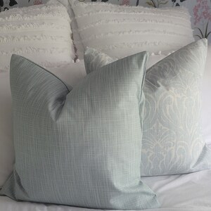 Puede incluir: Dos almohadas decorativas sobre una cama blanca. Una almohada es de color azul claro s&oacute;lido con una textura tejida sutil. La otra almohada es blanca con un patr&oacute;n floral azul claro.