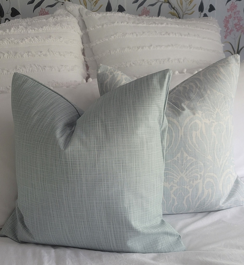 Puede incluir: Dos almohadas decorativas sobre una cama blanca. Una almohada es de color azul claro s&oacute;lido con una textura tejida sutil. La otra almohada es blanca con un patr&oacute;n floral azul claro.