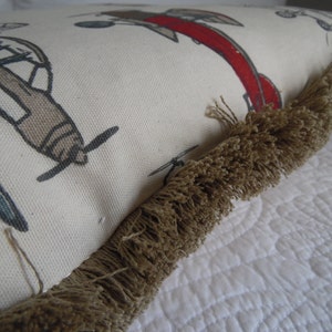 May include: A white pillow with a brown fringe border. The pillow has a pattern of airplanes and a red plane with a white outline.