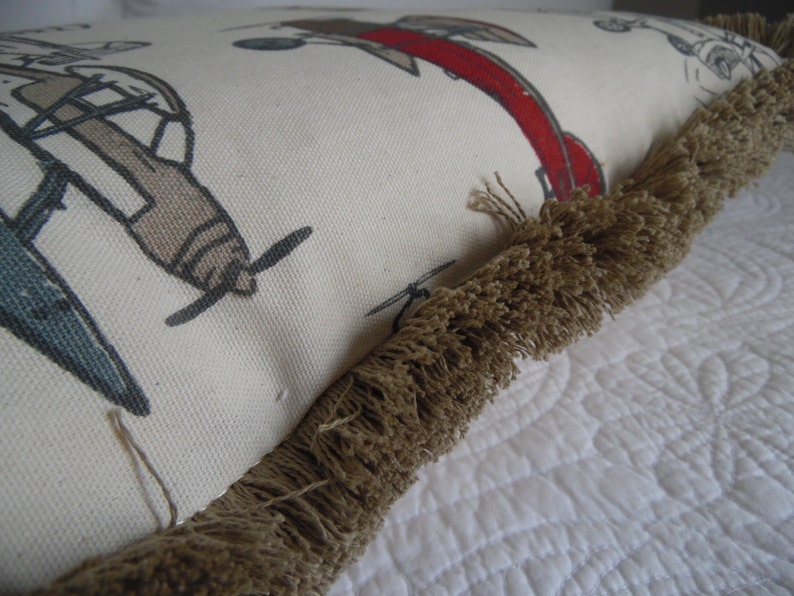 May include: A white pillow with a brown fringe border. The pillow has a pattern of airplanes and a red plane with a white outline.