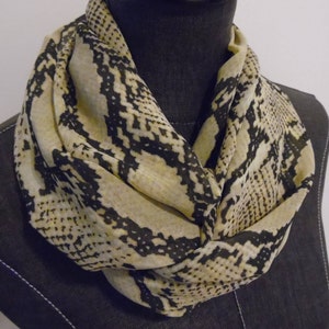 May include: A beige and black infinity scarf with a snakeskin pattern. The scarf is draped around a mannequin's neck, showcasing the fabric's drape and design. The pattern features dark black irregular shapes against a light beige background.