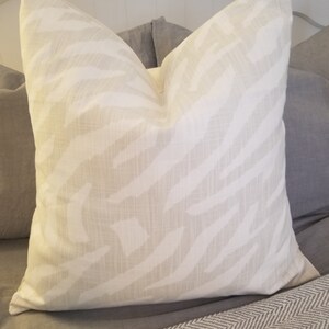 Puede incluir: Una almohada blanca con un sutil estampado de cebra beige. La almohada est&aacute; sobre una cama con una manta de patr&oacute;n de chevron gris y blanco.