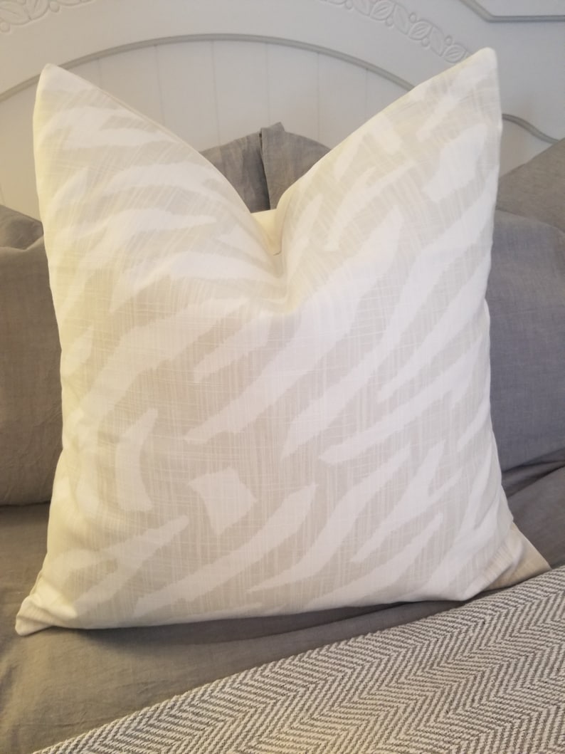 Puede incluir: Una almohada blanca con un sutil estampado de cebra beige. La almohada est&aacute; sobre una cama con una manta de patr&oacute;n de chevron gris y blanco.