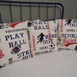 May include: Three white throw pillows with a sports theme. The pillows feature red, blue, and black text and graphics, including a football, baseball, and soccer ball. The text on the pillows includes "Play Ball", "Strike Out", and "Score Baseball".