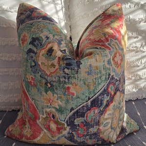 May include: Decorative throw pillow with a colorful, intricate pattern. The pillow features a mix of teal, red, yellow, and navy blue hues in a floral and paisley design. The pillow is placed on a blue surface with white dots and against white pillows.