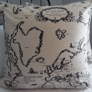 Può includere: Un cuscino beige con una mappa del mondo in bianco e nero stampata su di esso. La mappa include il testo "Map of the World" e "The world is a book and those who do not travel read only one page."