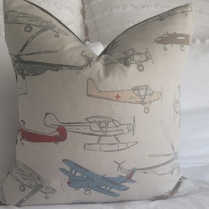 May include: A beige throw pillow with a brown trim features a repeating pattern of vintage airplanes in various colors and styles. The airplanes are drawn in a simple, graphic style and include a red biplane, a blue biplane, a yellow plane with a red cross, and a brown bomber.