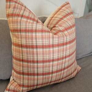 May include: A red, beige and white plaid pillow with a cream coloured border. The pillow is sitting on a grey bed.