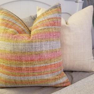 May include: A striped throw pillow with yellow, pink, and orange stripes on a gray background. The pillow is on a bed with a white headboard and a white and gray blanket.