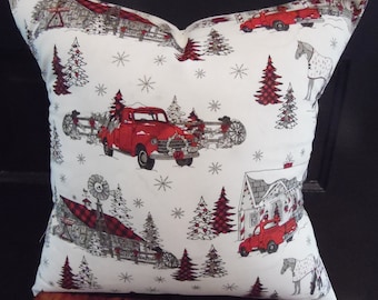May include: A square decorative pillow with a winter holiday theme. The white fabric is patterned with red trucks, barns, trees, and snowflakes. The pillow has a soft, inviting appearance, perfect for seasonal decor.