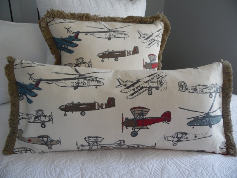 May include: Two decorative throw pillows with a fringe border. The pillows are made of a white fabric with a pattern of various airplanes in black, red, blue, and brown.