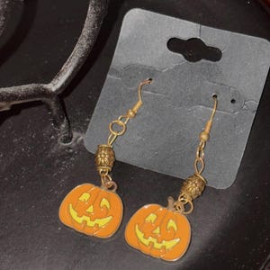 May include: A pair of gold dangle earrings with orange pumpkin charms. The pumpkins have a yellow smiling face with black eyes and a black mouth.