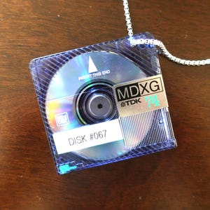 May include: A mini-disc necklace with a silver chain. The disc is in a clear plastic case. The disc has the text "INSERT THIS END" and "DISK #067". A gold sticker reads "MDXG TDK 74".